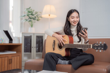 Young Asian woman sitting comfortably on a sofa in a cozy living room, playing acoustic guitar while reading chords on her smartphone, enjoying a relaxing practice session at home