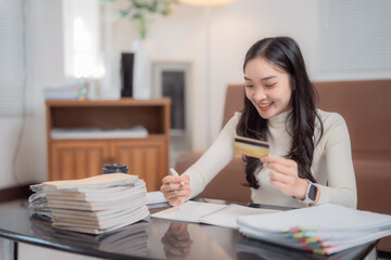 Young Asian businesswoman holding a credit card and jotting down notes while working from home,...