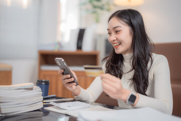Happy young Asian businesswoman holding a credit card and using her smartphone for online payments, enjoying the convenience of e-commerce and modern digital transactions