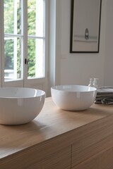 Fototapeta premium Two white sink sit atop a wooden surface near a window, complemented by neatly stacked towels and an artistic wall mirror in bathroom.