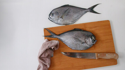 Two atlantic pomfret or Brama brama fish on a wooden cutting board with a knife and towel, set in a kitchen counter to show candid daily home life 