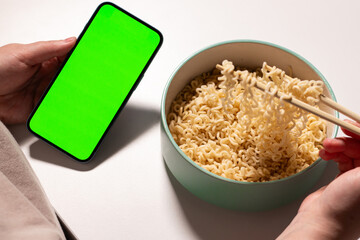 Female using smartphone while eating pasta at home. Smartphone with green screen with space for text. Smartphone in hands with served table in background