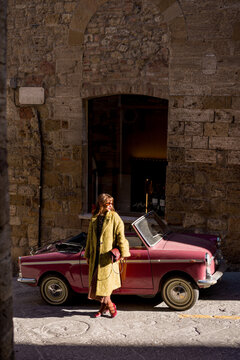 Fototapeta Stylish woman standing next to a classic convertible on a rustic street. A perfect combination of timeless fashion, Italian culture, and vintage charm