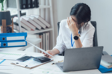 Overwhelmed by Work: A young businesswoman sits at her desk, head in her hands, surrounded by...