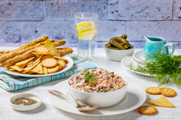 salmon dip with caper, red onion, fresh dill and sour cream in white bowl on white wooden table with crackers, tortilla chips and grissini in background of brick wall, horizontal view