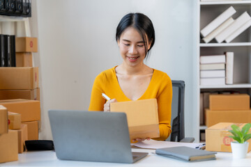 Online Business Owner: A cheerful, young Asian woman in a yellow sweater sits at her desk in a home...