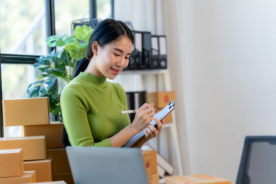 Focused on Fulfillment: A young entrepreneur meticulously checks her order list in a home office, a stack of cardboard boxes awaiting shipment in the background.