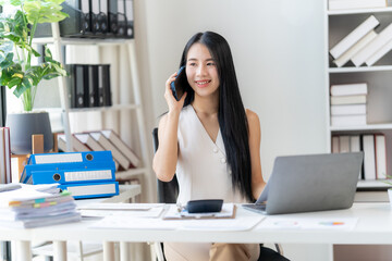 Confidently Connecting: A young Asian businesswoman smiles brightly as she conducts a phone call, demonstrating professionalism and efficiency at her desk with a laptop and documents.  