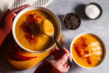 Woman tasting pumkin soup with spoon. Eating cream soup. High angle. Served table with soup bowl, pepper, chili, salt.