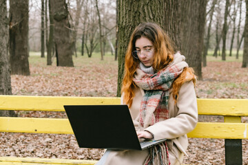 A young woman in glasses working on her laptop in a city park, writing or blogging outdoors. Ideal...