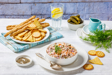 salmon salad with caper, red onion, fresh dill and sour cream in white bowl on white wooden table with crackers, tortilla chips and grissini in background of brick wall, horizontal view