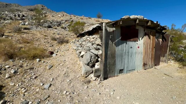 Rustic and weathered, a ramshackle miner's cabin constructed from local rocks and repurposed tin stands as a testament to pioneer ingenuity near Beatty, Nevada, capturing the essence of the region's r