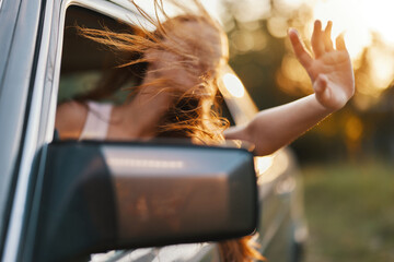 Young woman enjoying a summer drive with her hair blowing in the wind, wearing a casual outfit, expressing happiness with a joyful gesture outside the car window.