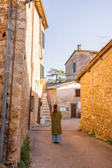 A woman in a green coat walks through a narrow village street surrounded by charming stone...