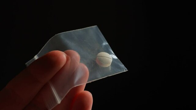 Hand of the drug dealer show off and lure with prescription opioid pill in small plastic bag for sale in clubs, parties and street to addicts. Illegal drugs trafficking, cause dependence and overdose