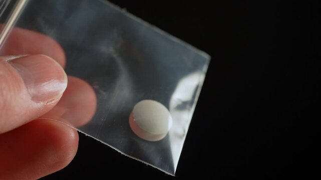 Hard drugs addictive person holding oxi opioid pill drug that he purchased from the dealer at the street. Close-up of a illegal drugs single pill in a small plastic bag held by hand, opioid crisis.