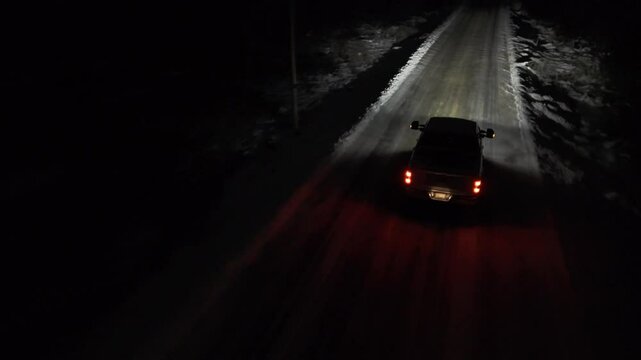 Aerial shots of a Pickup truck is driving down a snowy road at night. The headlights are on, illuminating the road ahead.