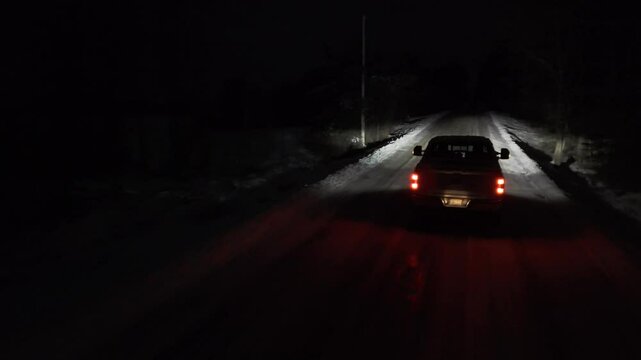 Aerial shots of a Pickup truck is driving down a snowy road at night. The headlights are on, illuminating the road ahead.