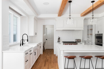 A white kitchen detail with a subway tile backsplash, large range hood, black faucet on a farmhouse sink and white marble countertop, and wood beams on the ceiling. No brands or labels.