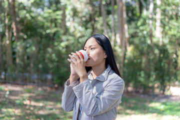 Fototapeta premium Drink coffee in the morning and prepare to welcome the new day. Asian woman stands relaxed in the middle of bright nature with sunlight shining on her face. Lifestyle concepts.
