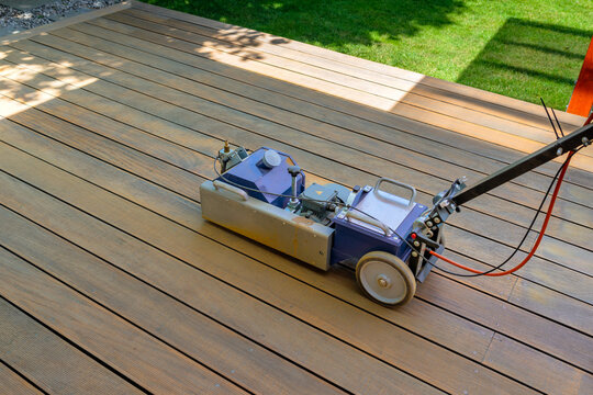 Worker cleaning and sanding a wooden deck with a specialized machine in a backyard setting with lush green lawn and a bright sunny environment.