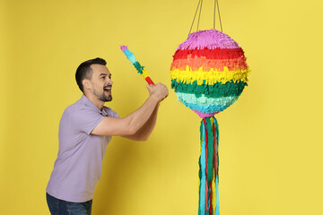 Emotional man hitting colorful pinata with stick on yellow background