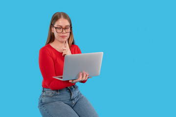 Young woman in red sweater using laptop while thinking in front of blue background
