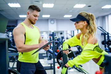 Obraz premium Fitness coaching in a bright gym setting. A fitness coach assists a female trainee in a well-lit gym, focusing on workout progress and motivation during training.
