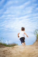 Child running on sandy path outdoors. A young child joyfully runs along a sandy path, surrounded by grass and a bright blue sky filled with clouds.