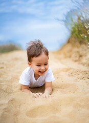 Child playing joyfully in sandy area. A young child lies on their stomach in the sand, smiling and enjoying the sunny outdoor environment near a path.