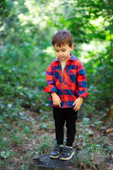 Fototapeta premium Kid on a log in the woods. A young child in a red and black checkered shirt stands on a log, surrounded by lush green trees and plants.