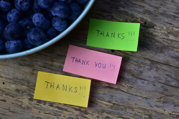 Message thanks thank you on pink green and yellow sticky notes day on wooden board textured background next to blueberries bowl