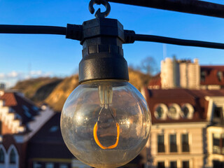 A vintage light bulb hangs from a bracket or hook in an urban setting, possibly outdoors The unlit...