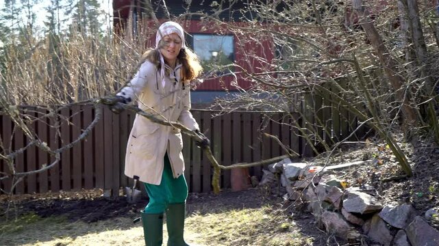 A woman dressed in outdoor workwear, gloves and a headscarf removes a tree branch in a garden during early spring clean up. The setting with rustic fence, a wooden house, and natural elements