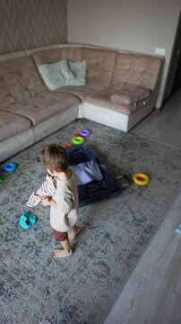 Small kid picks clothes from the floor scattered around the suitcase. Baby boy throws his things up with a joy. Top view. Vertical video.