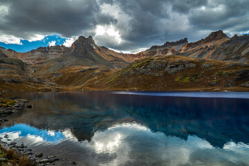 A stunning mountain lake mirrors rugged peaks and a vibrant, ever-changing sky in a serene and majestic natural landscape.