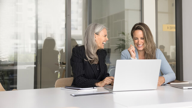 Two attractive businesswomen laughing during meeting in office, sitting at conference table with laptop, having fun, giggling, distracted from workflow, having pleasant, warm and friendly conversation