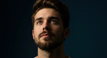 Obraz premium A charismatic young man with a stylish beard looks upward, with his features emphasized by dramatic lighting. 
