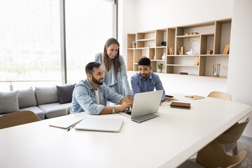 In modern conference room three multiethnic teammates engaged in workflow using laptop, discuss...