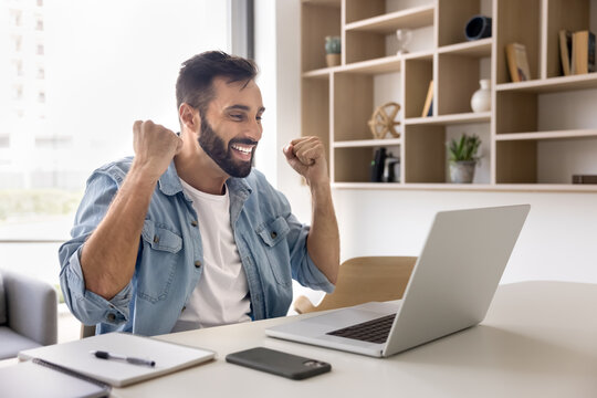 Young 30s Latin man sit at desk reading e-mail, clenching fists in celebratory yes gesture, get hired on job of dream, receive promotion, career growth opportunity, relish moment of auction victory