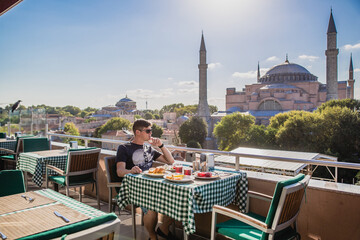Naklejka premium Young man enjoying breakfast on rooftop terrace with stunning view of Hagia Sophia in Istanbul, Turkey on sunny day