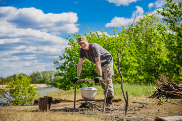 Man preparing fresh fish soup in pot and dry tree branches for campfire on river bank, resting...