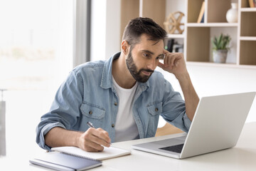 Young 30s Hispanic man sit at desk, gaze at laptop screen, jotting information, take notes,...