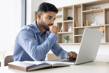 Focused man sit at desk reviewing data, analyzing reports, working on presentation or project plan, brainstorm ideas, doing telework, ponder, planning engaged in analytical tasks using modern laptop