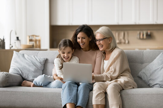 Funny movie. Friendly family of 3 generation mid aged granny young mother cute little tween age daughter grandchild spend free time hugging on sofa using notebook make shopping online watch video blog