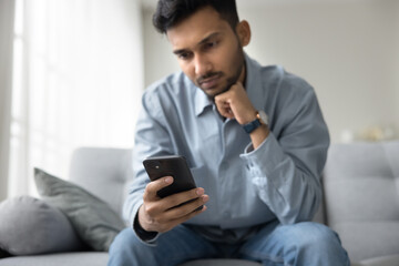 Serious millennial male sit on sofa at home read news from smartphone screen engaged in virtual communication scroll web pages use e-commerce application. Focus on modern cell device in young man hand
