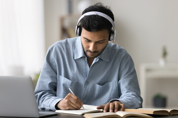 Effective online education. Focused smart gen z male remote student in wireless earphone device listen to virtual learning event web lecture distant course via notebook pc write in copybook take notes