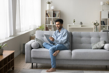 Obraz premium Digital leisure. Happy handsome millennial male relax on sofa alone at modern living room interior hold tablet computer device text with girlfriend in chat play online game download e-book. Copy space