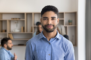 Head shot portrait young male office employee, Arabic businessman dressed in casual shirt posing for camera exuding professionalism and competence standing at workplace with colleagues on background