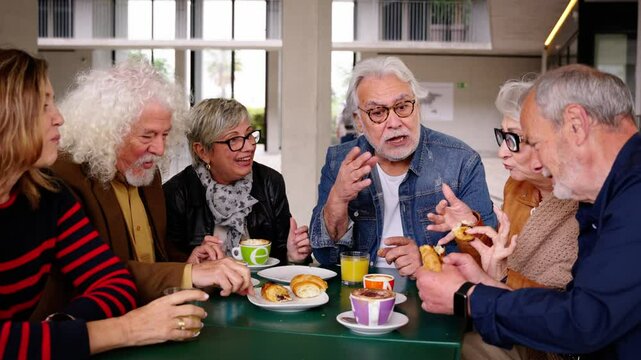 Group of joyful elderly friends gathered on terrace of residence cafeteria eating and drinking coffees and pastries. Caucasian seniors people meeting enjoying snack sitting together at table